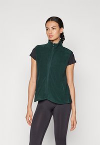 Chaleco de forro polar verde con cuello alto, cierre frontal de cremallera y bolsillos laterales; llevado sobre una camiseta negra ajustada. Textura suave, diseño casual.