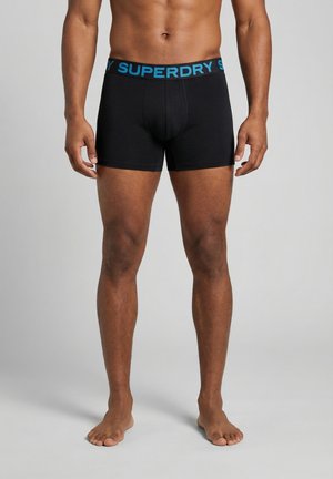 Mannelijk model draagt zwarte boxers met een blauwe "SUPERDRY" tailleband, staat blootsvoets tegen een effen grijze achtergrond.