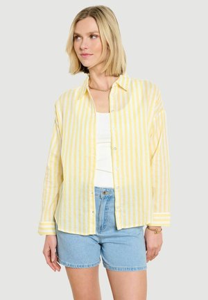 Frau mit kurzen blonden Haaren, die ein gelb-weiß gestreiftes Hemd über einem weißen Oberteil und hellblauen Jeansshorts trägt.