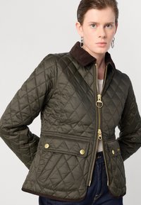 Gequilted olijfgroene jas met een bruine corduroy kraag, gouden rits en voorkruiszakken met knoopsluitingen. Gelaagd stof en op maat gemaakte pasvorm.