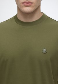 Gros plan d’une personne portant un t-shirt vert olive à col rond avec un petit logo rond sur la poitrine gauche affichant le chiffre huit.