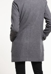 Cappotto grigio in lana con una vestibilità sartoriale, rever a punta e dettagli di cucitura sottili; presenta maniche lunghe e un taglio di lunghezza media.