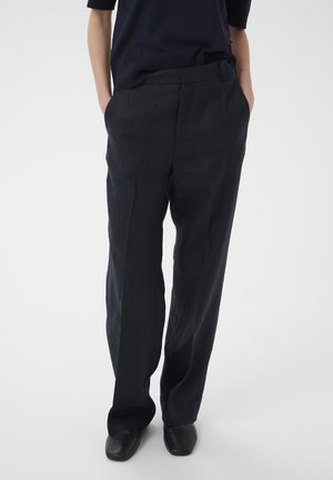 Osoba oblečená v čiernych nohaviciach s širokými nohavicami, ruky vreckách, v kombinácii s čiernou obuvou a tmavým vrchom na jednofarebnom pozadí.