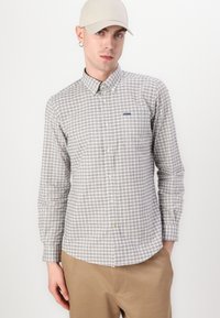 Chemise à carreaux en beige et gris, avec des manches longues, un col et un petit logo de marque sur le côté gauche.