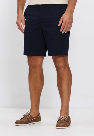 Uomo con pantaloncini blu navy e scarpe da barca marroni, in piedi contro uno sfondo chiaro e uniforme.