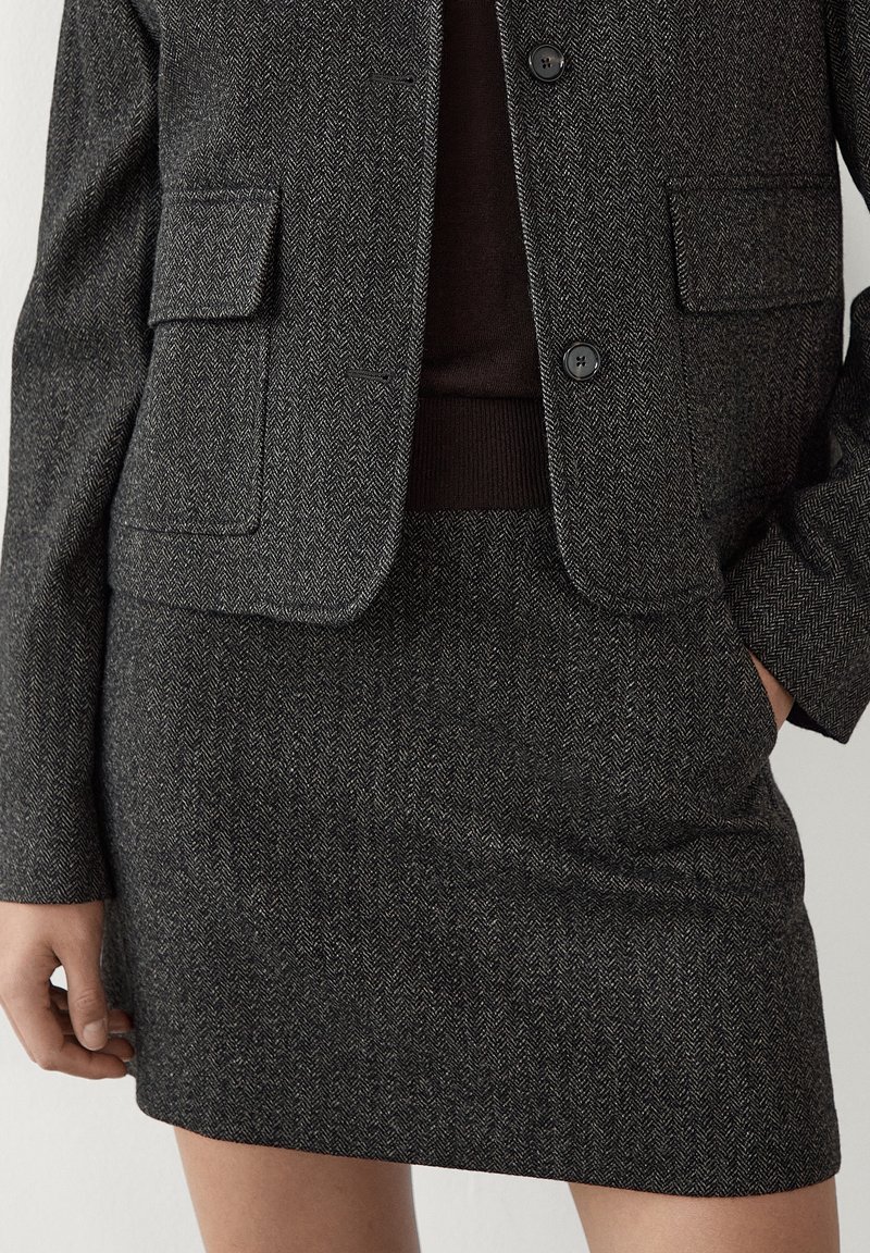 Houndstooth-patterned, dark gray blazer with two front pockets and a fitted skirt. Fabric features a textured finish with subtle sheen.