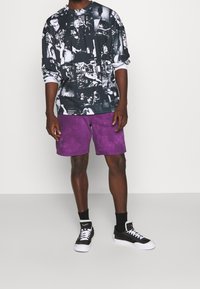 T-shirt graphique noir et blanc à manches longues avec une coupe décontractée, associée à un short violet tie-dye et des baskets noires avec des semelles blanches.
