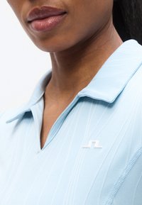 Ανοιχτό μπλε πόλο πουκάμισο με υφή σχεδίου. Διαθέτει μυτερό γιακά και λευκή λογότυπο στο στήθος. Ομαλή υφή υφάσματος.