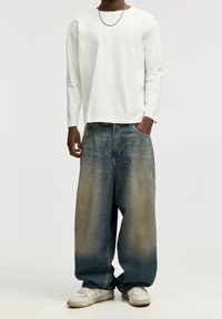 Camisa blanca de manga larga, corte holgado, combinada con jeans baggy azul claro descoloridos hacia un tono más claro en la parte inferior. Zapatillas deportivas blancas.