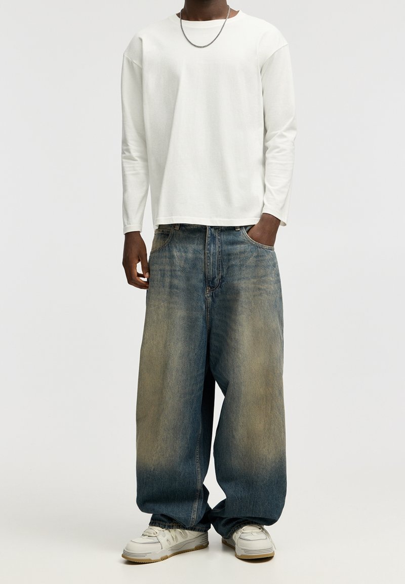 Camisa blanca de manga larga, corte holgado, combinada con jeans baggy azul claro descoloridos hacia un tono más claro en la parte inferior. Zapatillas deportivas blancas.