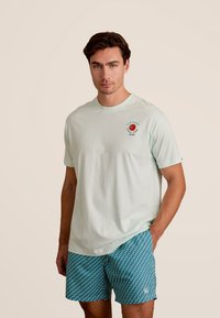 Licht mintgroene katoenen T-shirt met korte mouwen, met een cirkelvormig rood en blauw logo op de borst. Gecombineerd met gestreepte blauwe shorts.