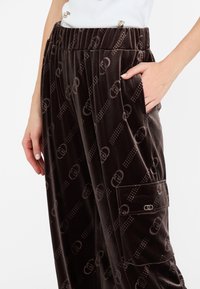 Pantaloni cargo in velluto marrone con una texture morbida, caratterizzati da un motivo a logo ripetuto, vita elastica e dettagli con tasche laterali.