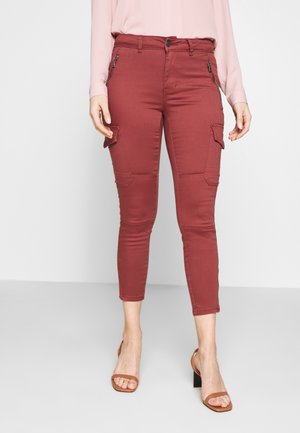 Kobieta nosząca wysokie, krótkie rdzawoczerwone spodnie cargo z zamkami i kieszeniami z klapami, w połączeniu z brązowymi sandałami na obcasie z odkrytymi palcami.