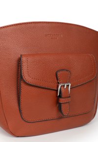 Borsa a mano in pelle marrone con una texture liscia, dotata di una tasca frontale fissata da una fibbia argentata. Forma arrotondata, design minimale, dettaglio del marchio.