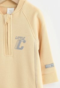 Helle beigen Baby-Hoodie aus weichem Stoff, mit einem halben Reißverschluss, gerippten Bündchen und grauem Drucktext "KLEINER BÄR" sowie Logo.