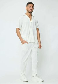Chemise blanche à manches courtes et boutons avec un motif texturé en vagues, assortie à un pantalon fuselé, portée avec des baskets blanches.
