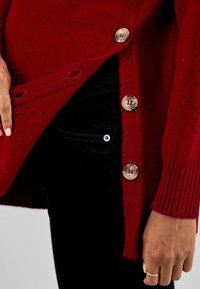 Cardigan rojo de punto con mangas acanaladas y grandes botones brillantes, combinado con pantalones negros que muestran un detalle visible de bolsillo lateral.