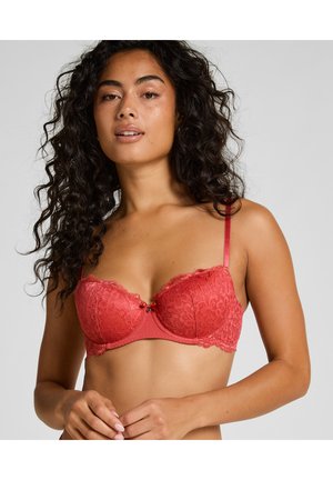 Frau mit lockigem Haar, die einen roten Spitzen-Balconette-BH trägt, vor einem schlichten hellgrauen Hintergrund stehend.