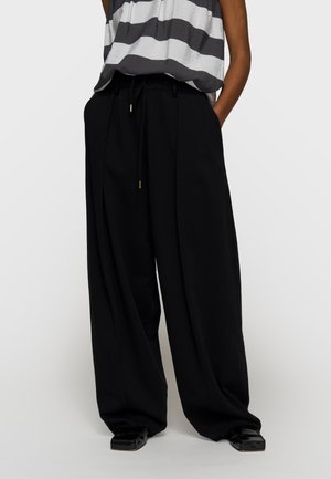 Modella che indossa pantaloni neri larghi e morbidi con vita regolabile a coulisse e scarpe nere con punta quadrata, abbinati a una maglia a righe grigie e bianche.