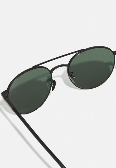Round black sunglasses with dark green lenses, thin black metal frame, and sleek design. Features nose pads for comfort and simple accents.