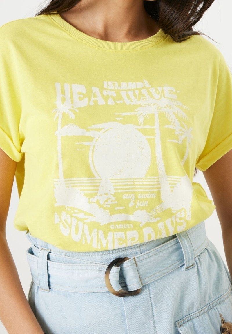 Kvinna iklädd en gul t-shirt med vit tropisk tryck som står "Island Heat-Wave Summer Days", instoppad i högmidjade ljusblå denimbyxor.