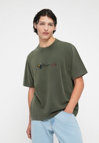 Olivgrön bomulls T-shirt med korta ärmar, prydd med en broderad design av en kajak och paddel, i kombination med ljusblå jeans.