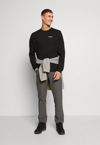 Sort langærmet skjorte med lille logo, grå bukser og sorte støvler. Grå sweatshirt bundet om taljen med gule detaljer på ærmerne.