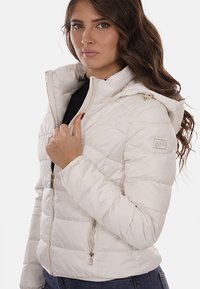 Giacca a vento bianca con chiusura a zip, cappuccio e design trapuntato. Presenta dettagli in metallo dorato e tasche laterali. Texture liscia.