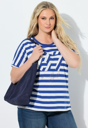 Blonde Frau in blau-weiß gestreiftem Hemd mit dem Text "87 sunlight", die eine marineblaue Umhängetasche hält und vor einer weißen Wand posiert.