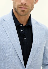 Primo piano di un uomo che indossa un blazer a spina di pesce blu chiaro su una polo a maglia nera con bottoni, che mostra una leggera barba e labbra neutre.