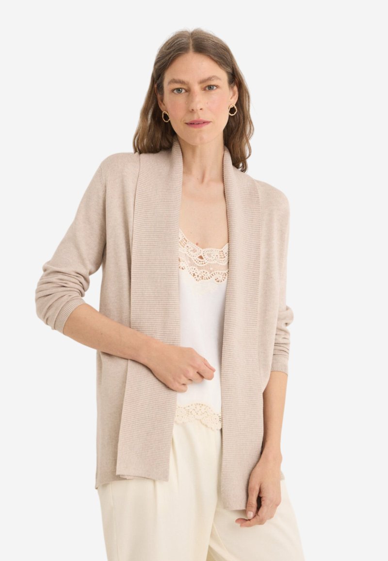 Cardigan beige con colletto sciallato a costine, apertura frontale e maniche lunghe. Indossato sopra una maglietta bianca con rifiniture in pizzo. Tessuto morbido e texturizzato.