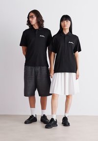 Camisas polo negras con un logo blanco, combinadas con pantalones cortos grises y una falda plisada blanca; ambos modelos llevan zapatillas oscuras y calcetines hasta la pantorrilla.