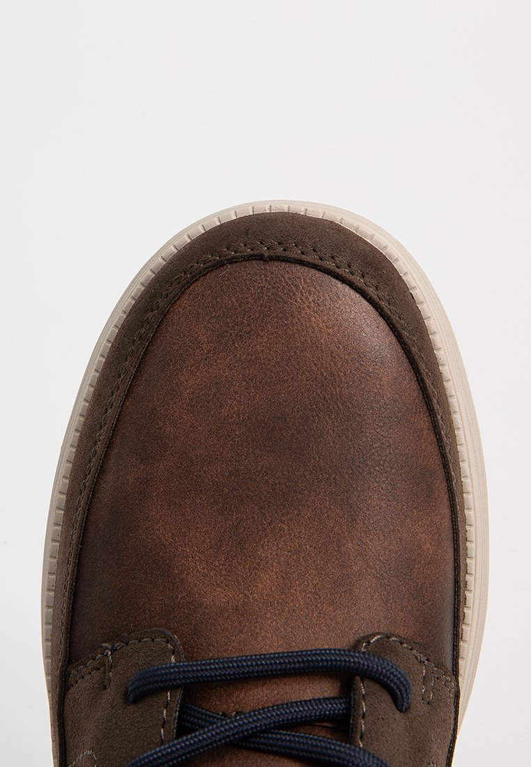 Zapatilla de cuero marrón con puntera redondeada, superficie texturizada y cordones en azul oscuro en contraste, con una suela de goma blanca.