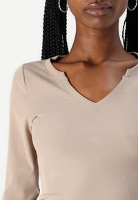 Beige színű, hosszú ujjú, bordázott anyagú felső, V-nyakú kivágással és testhezálló fazonnal. Megjegyzésre méltó varrás a kivágás körül.