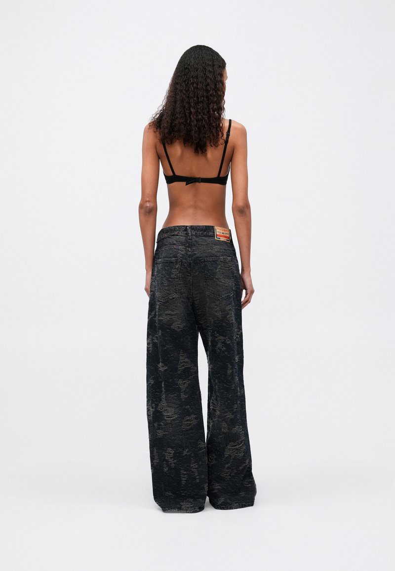 Frau mit lockigem Haar, die sich abkehrt und einen schwarzen BH sowie weite, schwarze Jeans mit einem Patchlabel am hinteren Bund trägt.