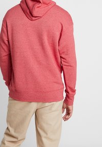 Röd huvtröja i en strukturerad textur, med dragsko i huvan och ribbade ärmslut, kombinerad med beige byxor.