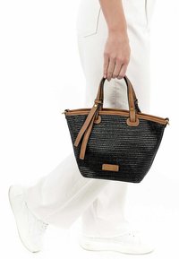 Persoon in witte broek en sneakers, die een kleine zwarte geweven handtas vasthoudt met bruin leren handvatten en afwerkingen.