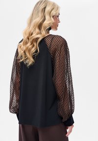 Czarna bluzka z przezroczystymi, brązowymi rękawami w kropki; cechuje ją luźny krój i gładka faktura, z długimi rękawami i okrągłym dekoltem.