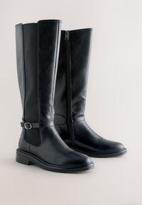 Paar hohe schwarze Lederstiefel mit seitlichem Reißverschluss und dekorativem Riemen mit Schnalle auf neutralem Hintergrund.