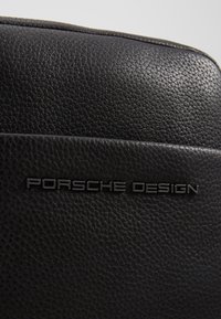 Czarna torba z fakturowanej skóry z wytłoczonym logo „Porsche Design” w eleganckim metalicznym kroju na gładkiej panelu.
