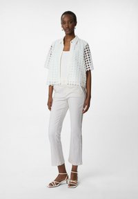 Biała koronkowa kurtka z geometrycznymi wycięciami, w połączeniu z białą bluzką i jasnymi, prostymi spodniami. Uzupełniona o sandały na paski.