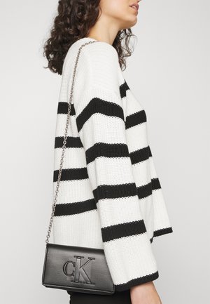Mujer con un suéter blanco con rayas horizontales negras, llevando un bolso de cuero negro Calvin Klein con una correa de cadena plateada en el hombro.