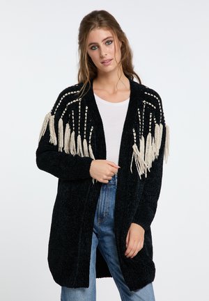 Junge Frau trägt eine schwarze Strickjacke mit weißen Fransen über einem weißen Hemd und blauen Jeans, steht vor einem weißen Hintergrund.
