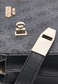 Svart texturerad handväska med guldfärgad hårdvara, ett rektangulärt spänne och en cirkulär låsdetalj. Materialet verkar vara syntetiskt och hållbart.