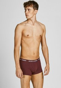Young man with short brown hair wearing maroon JACK & JONES boxer briefs, looking left against a plain gray background.