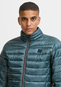 Teal pufferjack met een hoge kraag, doorgestikte ontwerp, zichtbare stiksels en een contrasterende rits. Heeft een klein logo op de borst.