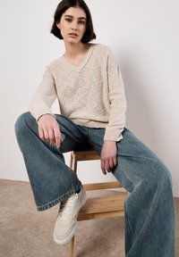Beige gebreide trui met een V-hals, gecombineerd met een wijdvallende spijkerbroek in denimblauw. Het model draagt lichtgekleurde sneakers en zit op een kruk.