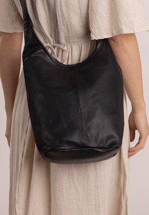 Czarna skórzana torba na ramię o miękkiej teksturze, zaokrąglonym kształcie i szerokim pasku, oparta o jasnokolorową tkaninę.