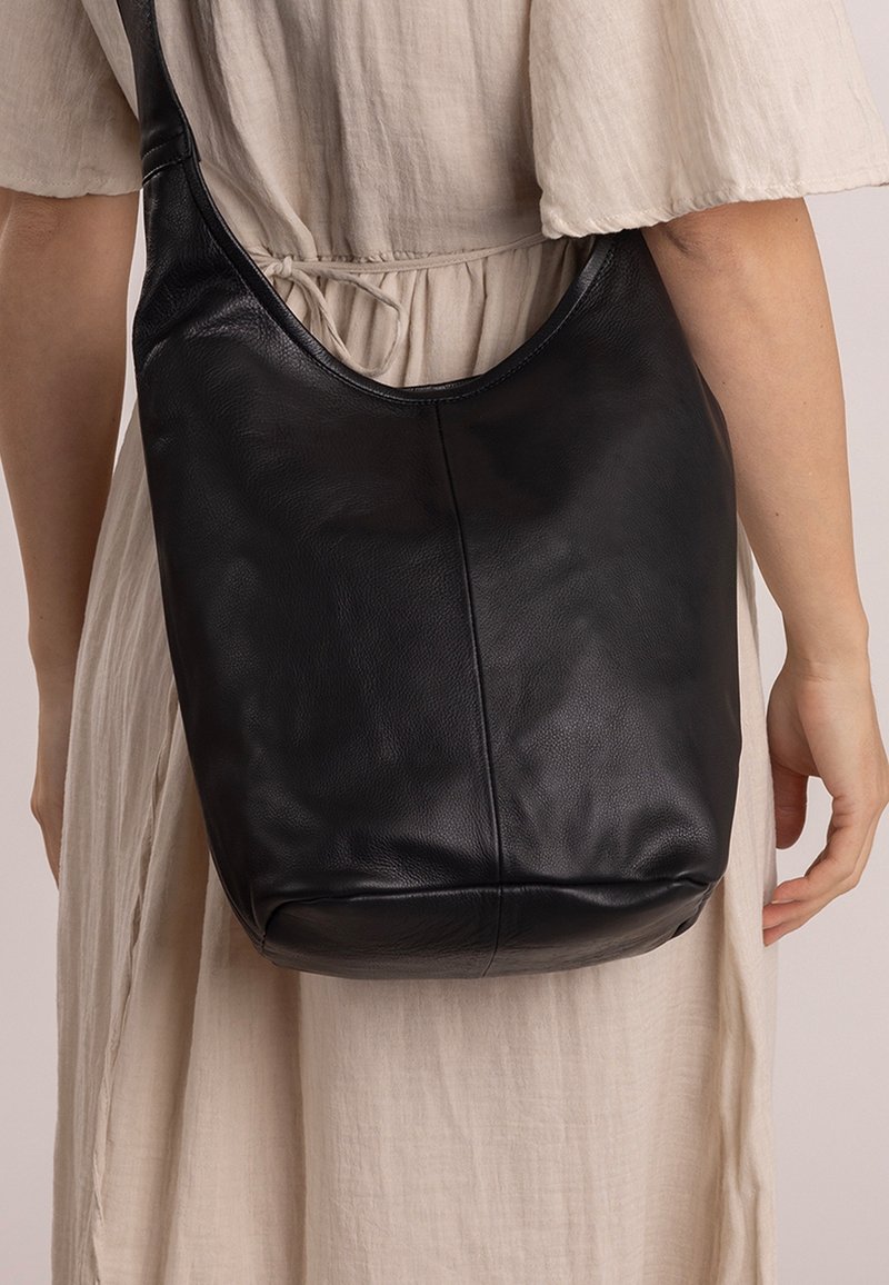 Borsa a tracolla in pelle nera dalla texture morbida, forma arrotondata e tracolla larga, appoggiata su un tessuto di colore chiaro.