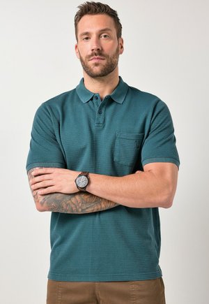 Mężczyzna z brodą i tatuażami w turkusowej koszulce polo i brązowych spodniach, z założonymi ramionami, o neutralnym wyrazie twarzy, na jasnym, jednolitym tle.